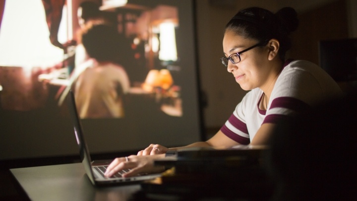 student working on computer with film showing in background