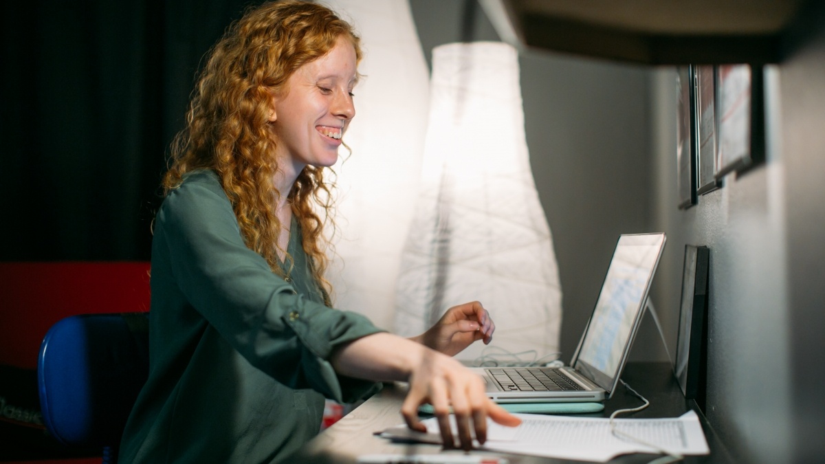 red headed person smiling while looking at a screen and grabbing for papers