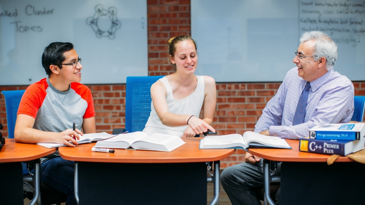 Professor and two students sitting at a table with books in discussion.